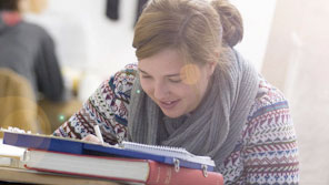 Student writing notes on a pile of books in a study area